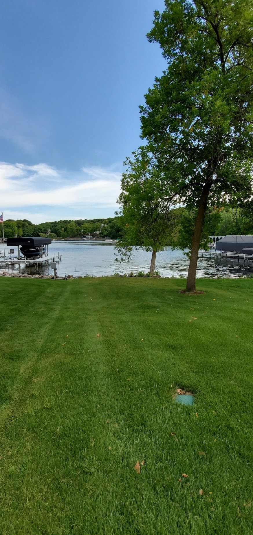 Green grassy field leads to a lake with boats, under a blue sky with trees.