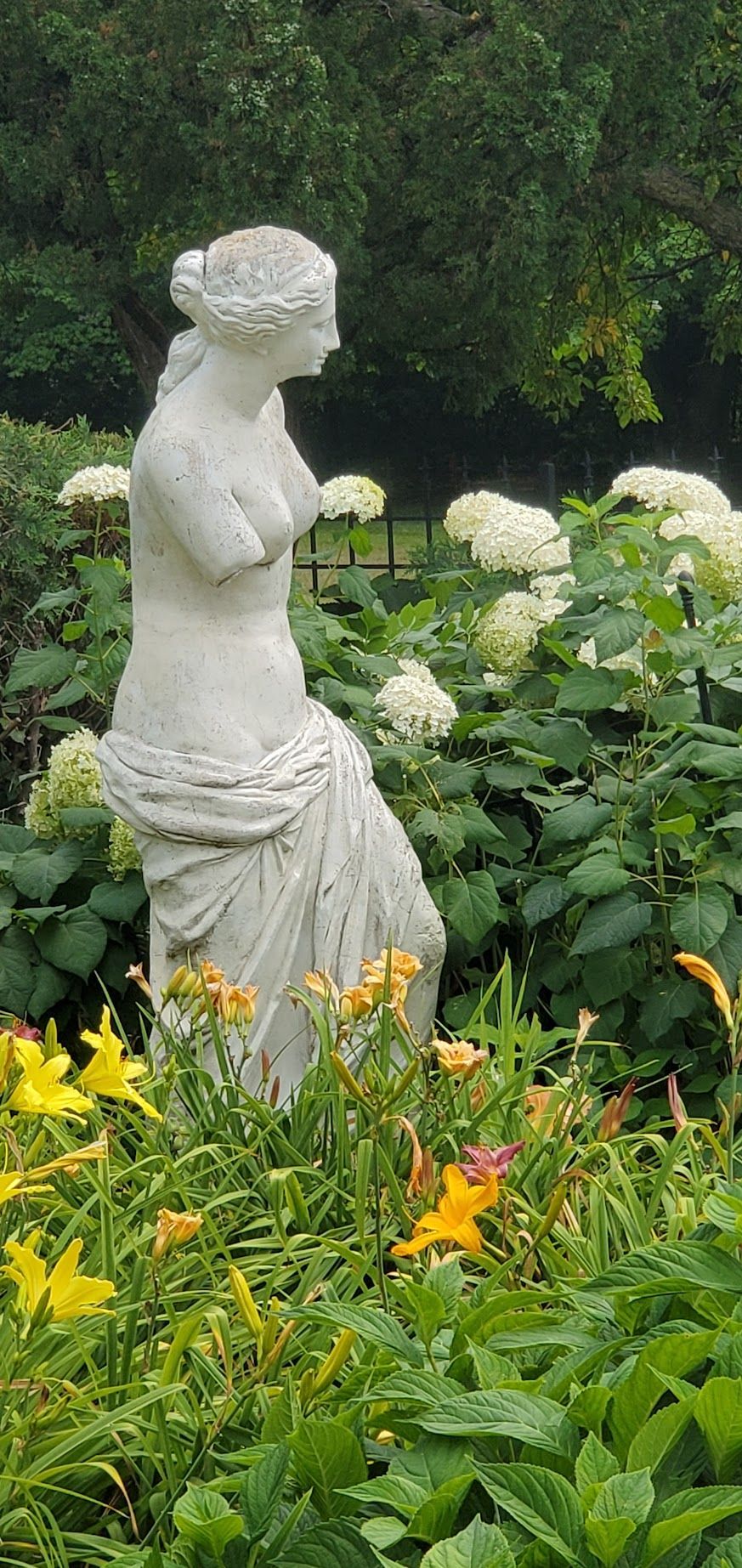 Statue of a woman in a garden, with flowers and greenery.