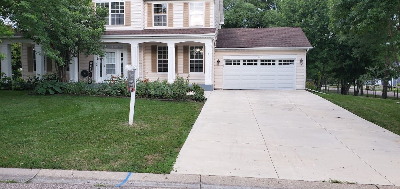Two-story house with white trim, tan siding, and a concrete driveway. Green lawn in front.