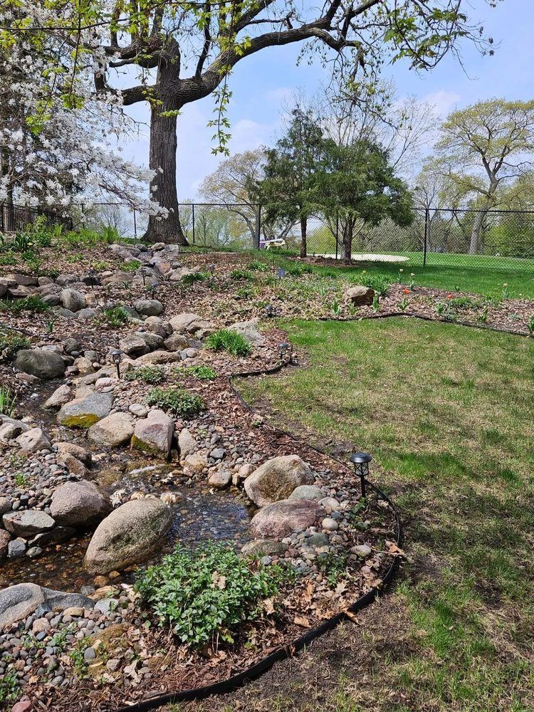 Stone-lined stream flows through a landscaped garden with a large tree. Green grass and blue sky in the background.