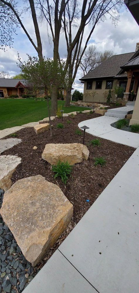 Landscaped yard with large rocks, mulch, and a concrete path leading to a house.