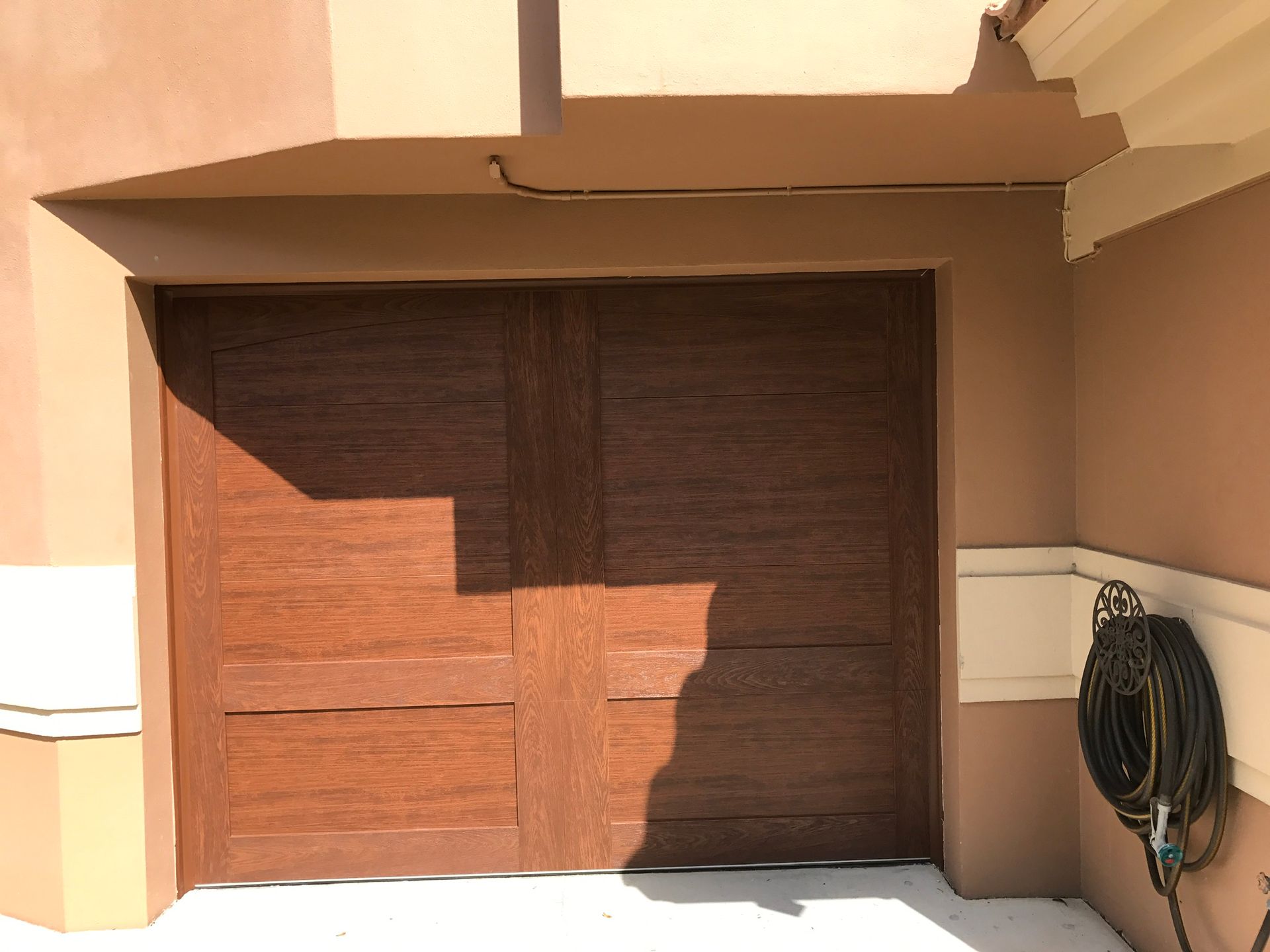 Wooden garage door with a hose, set in a beige and brown building. Sunlight casts shadows.
