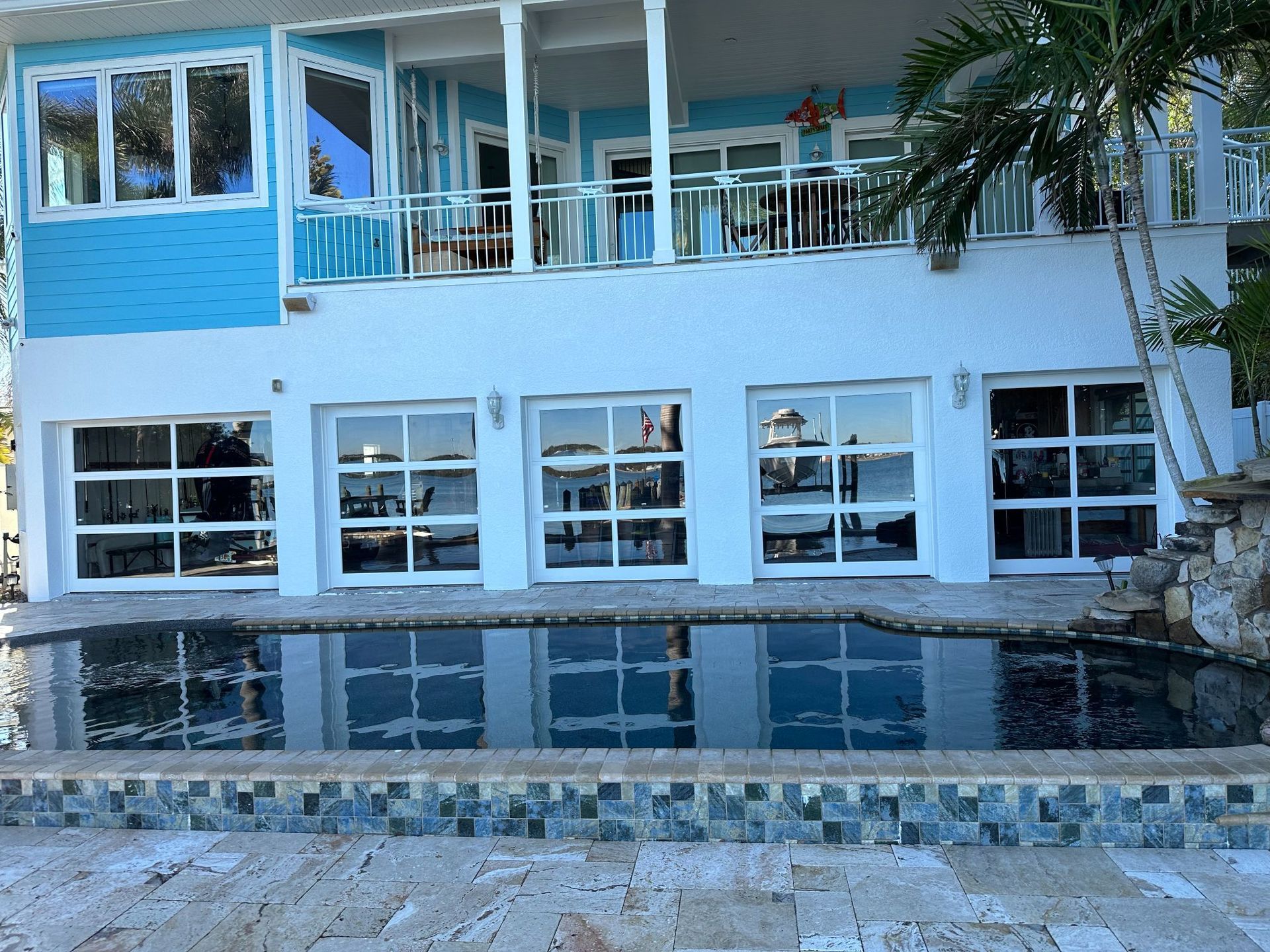 Bright blue waterfront house with pool, clear garage-style doors, balcony, and reflections in the water.