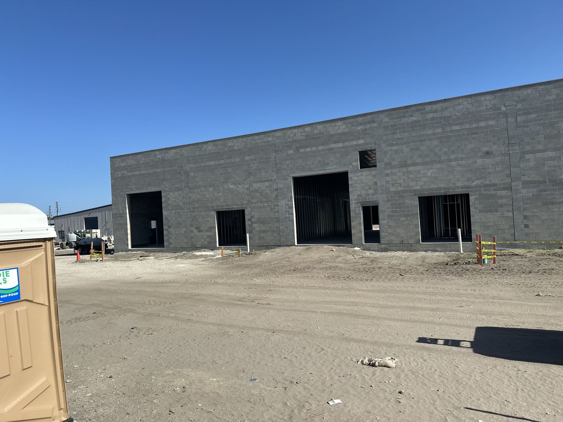 A building under construction with large open doorways, a portable toilet in the foreground, and a blue sky.