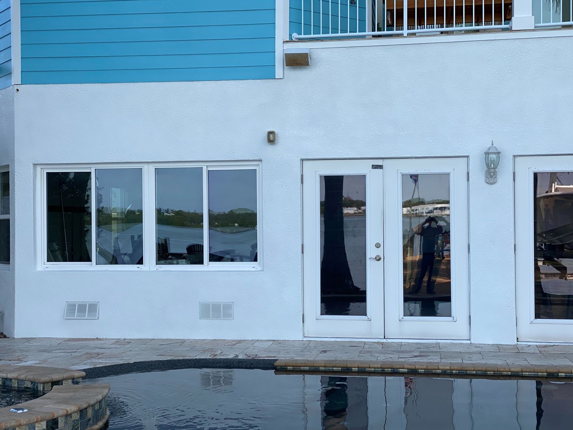 White house with pool, windows, and glass doors reflecting a person, a body of water, and a balcony.