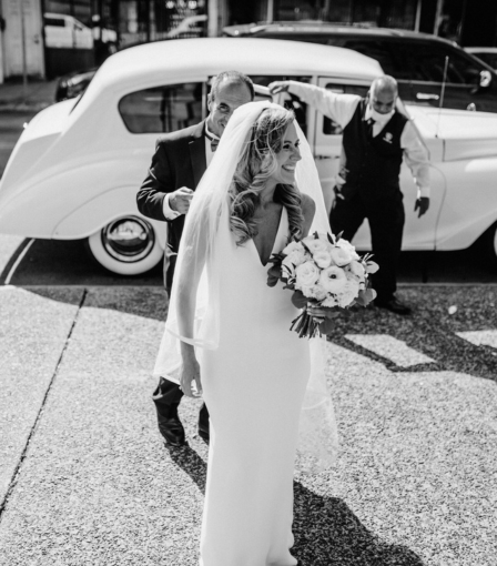 A black and white photo of a bride getting out of a white car