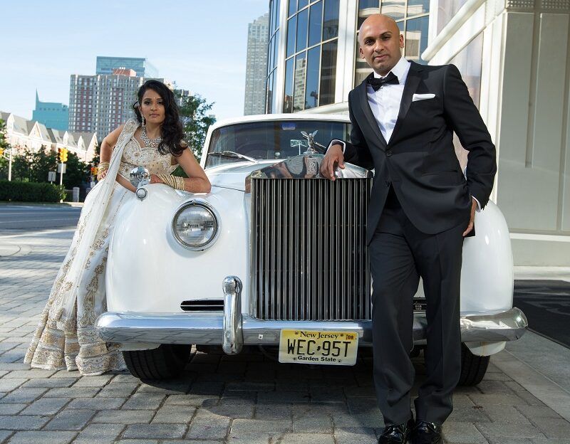 A bride and groom pose in front of a white rolls royce