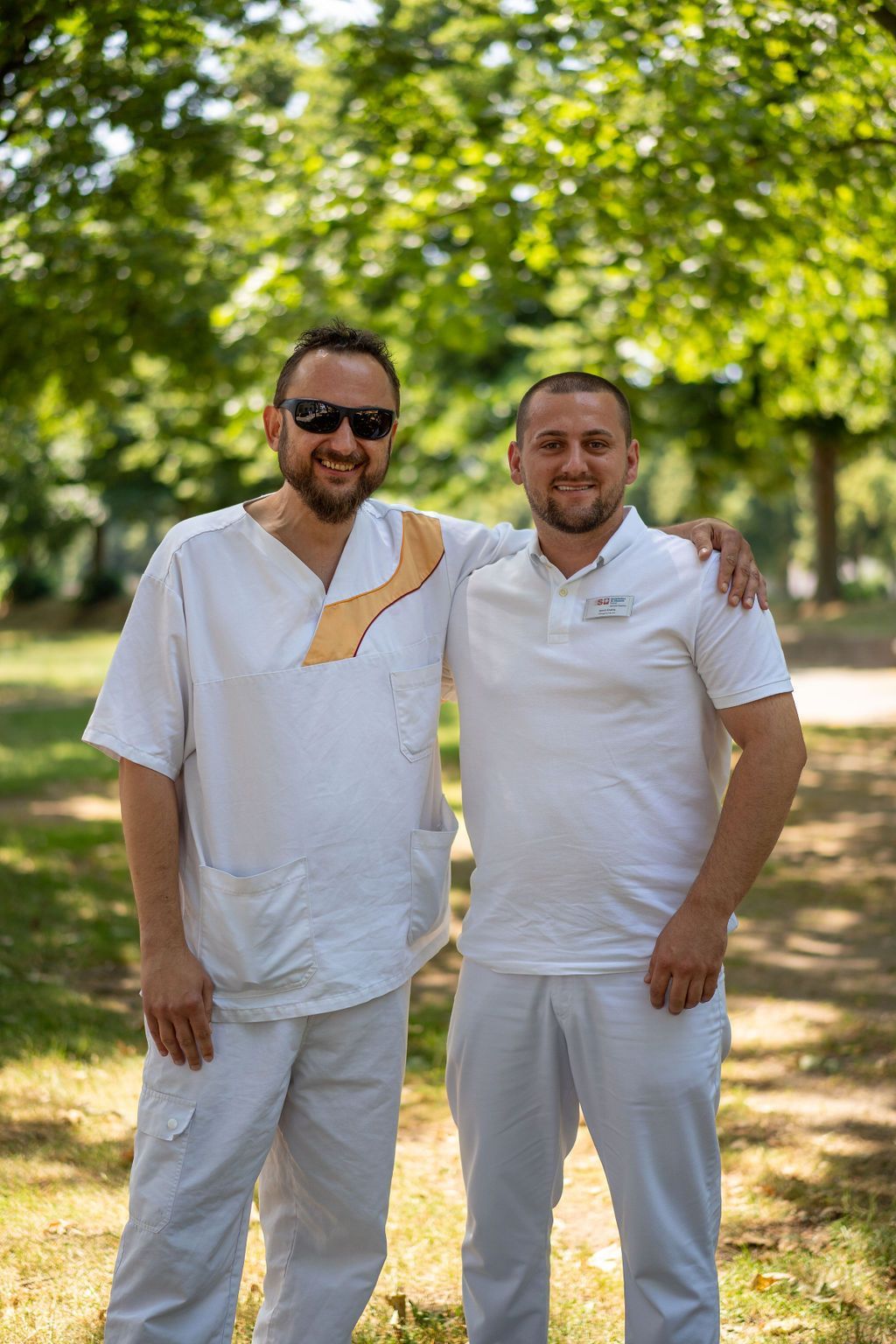 Zwei Pflegefachmänner in Dienstkleidung Arm in Arm im Garten