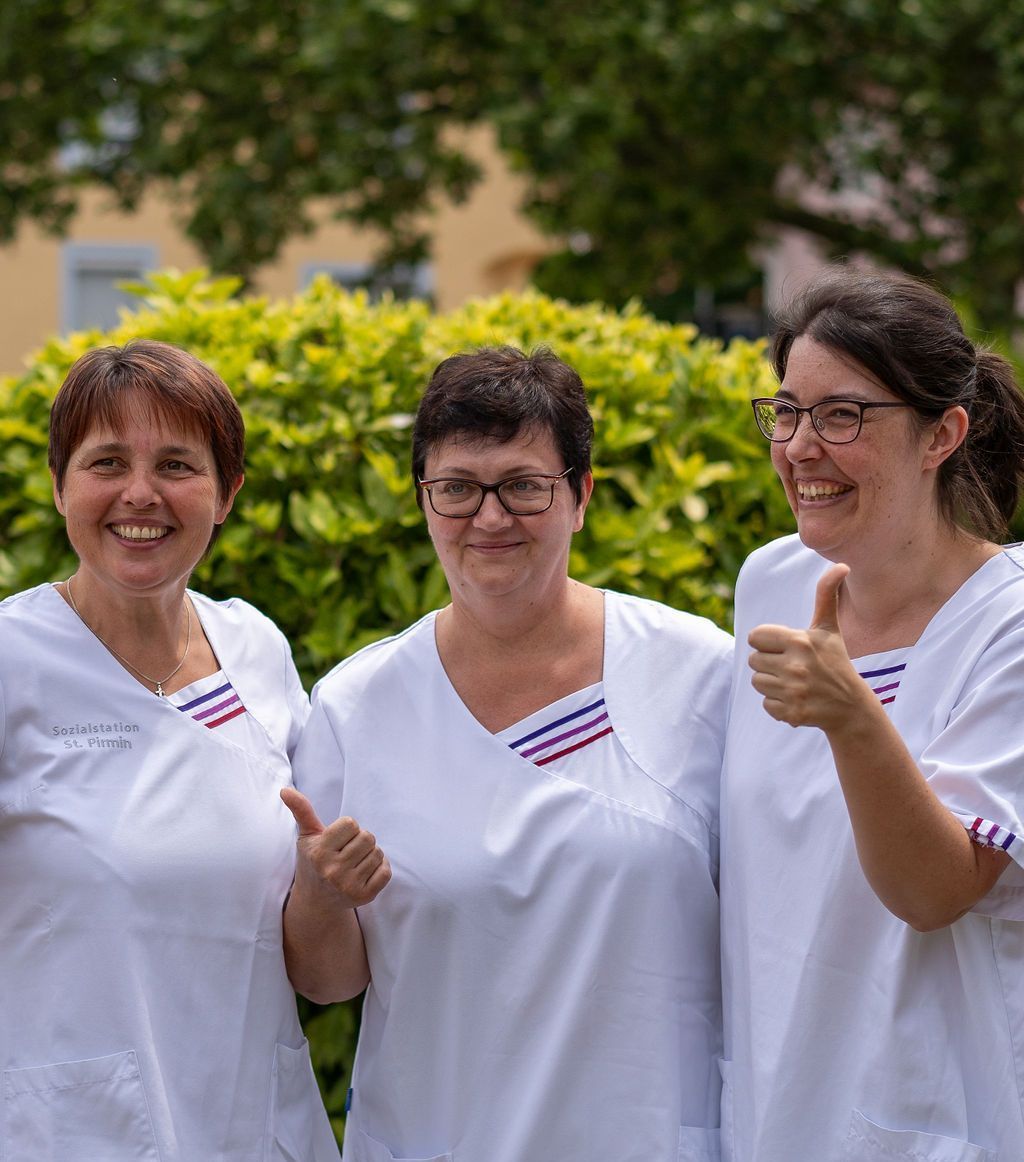 Drei Pflegefachkräfte Arm in Arm mit Daumen hoch in Dienstkleidung