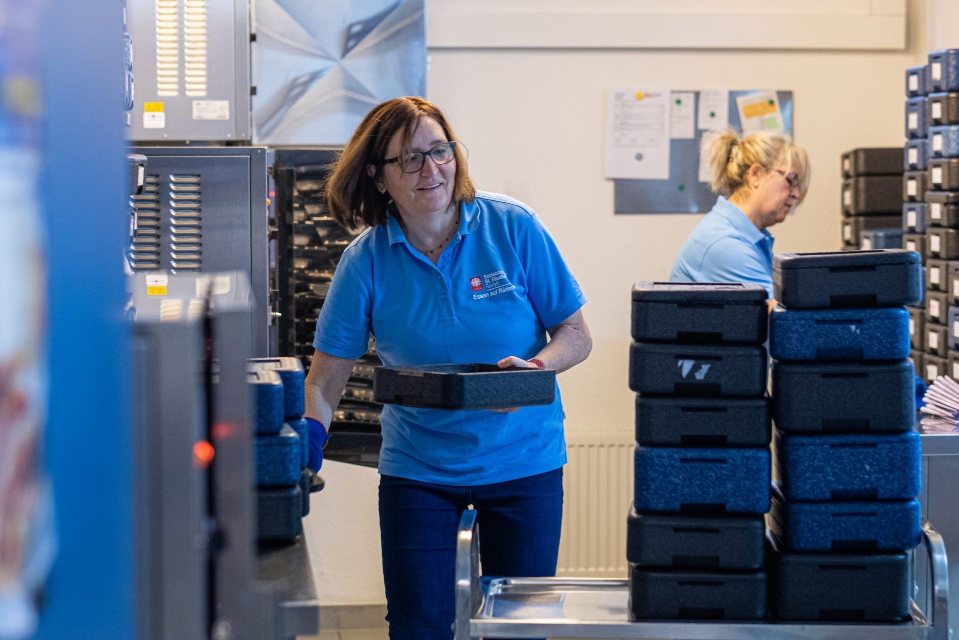 Mitarbeiterinnen bereiten Mahlzeiten in Boxen für Essen auf Rädern vor.
