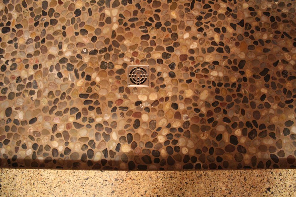 Shower floor with dark, rounded river stones surrounding a drain. The bottom border is tan.