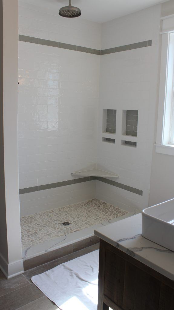 White tiled shower with grey accents, two built-in shelves, and a shower head, near a window and vanity.
