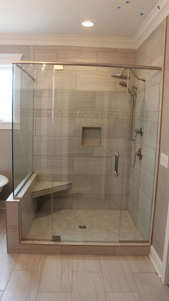 Glass-enclosed shower with built-in bench, grey tile, and silver fixtures, in a bathroom with beige flooring and walls.