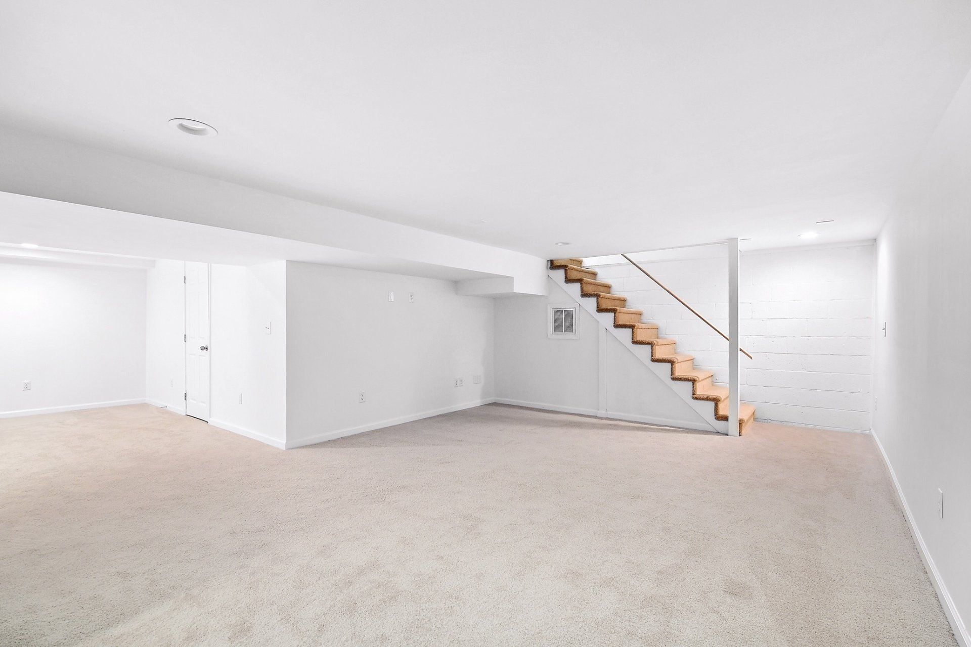 Empty basement with beige carpet, white walls, and wooden stairs.