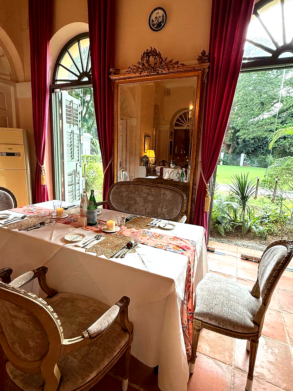 A dining table set at Suffolk House, for four in a room with arched doorways, deep pink curtains, a large ornate mirror, and garden views.