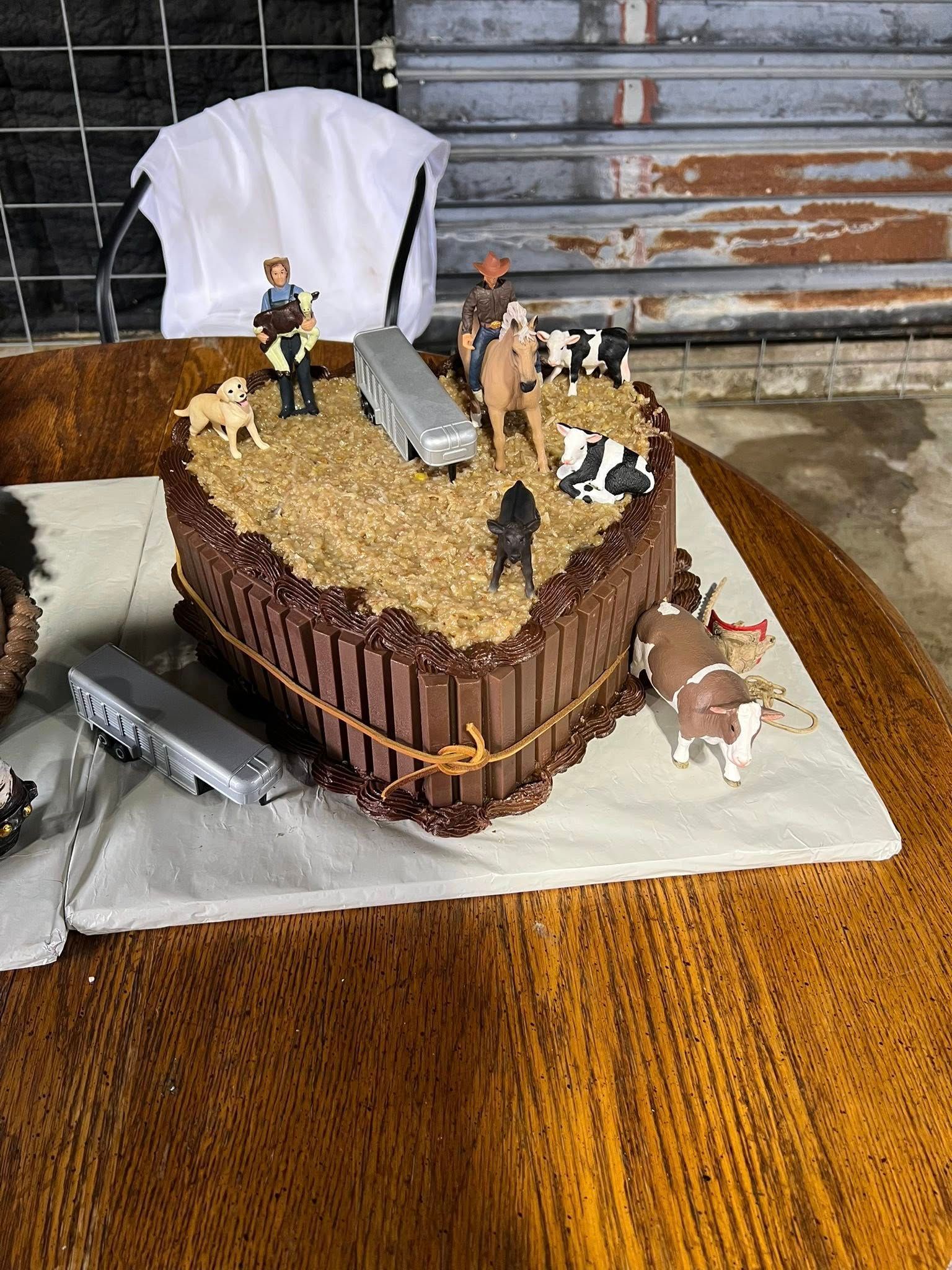 Heart-shaped cake decorated with a farm scene; toy figures of cows, a horse, and people on brown table.
