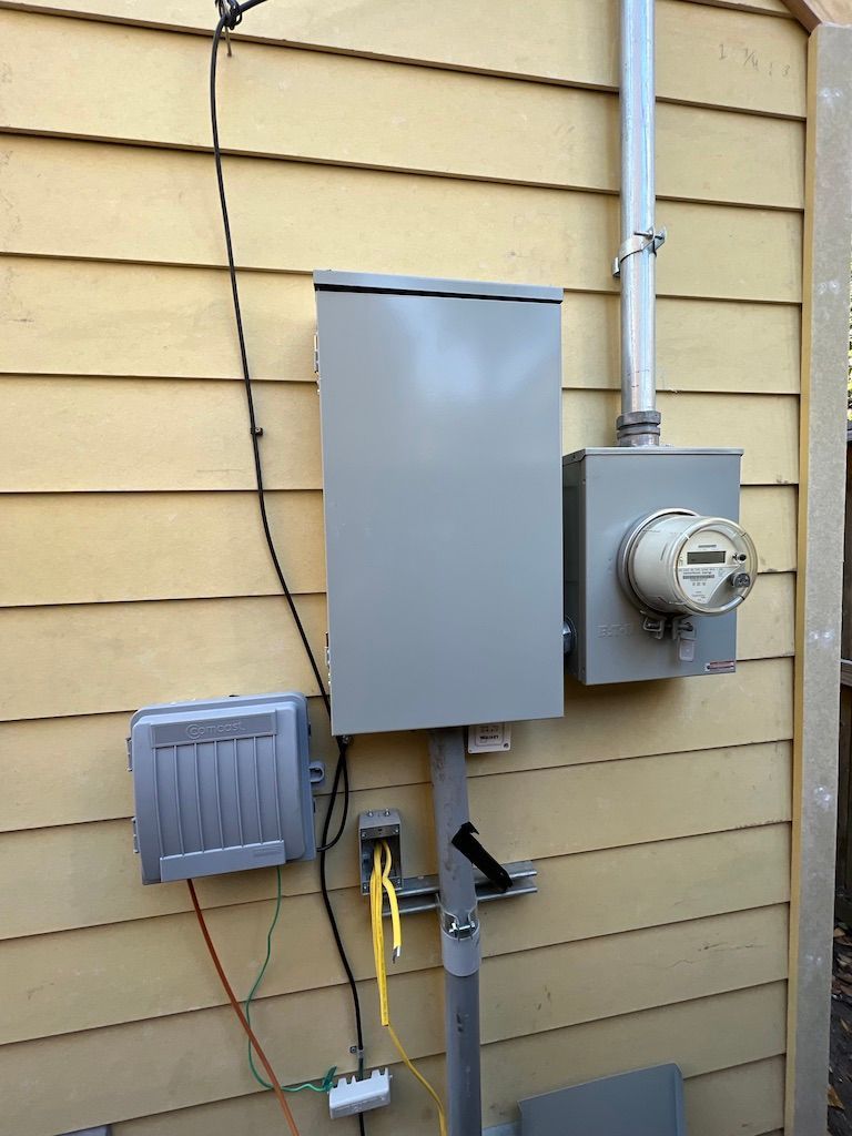 Electrical meter box and breaker panel mounted on a yellow-sided building. Utility lines and conduit are visible.