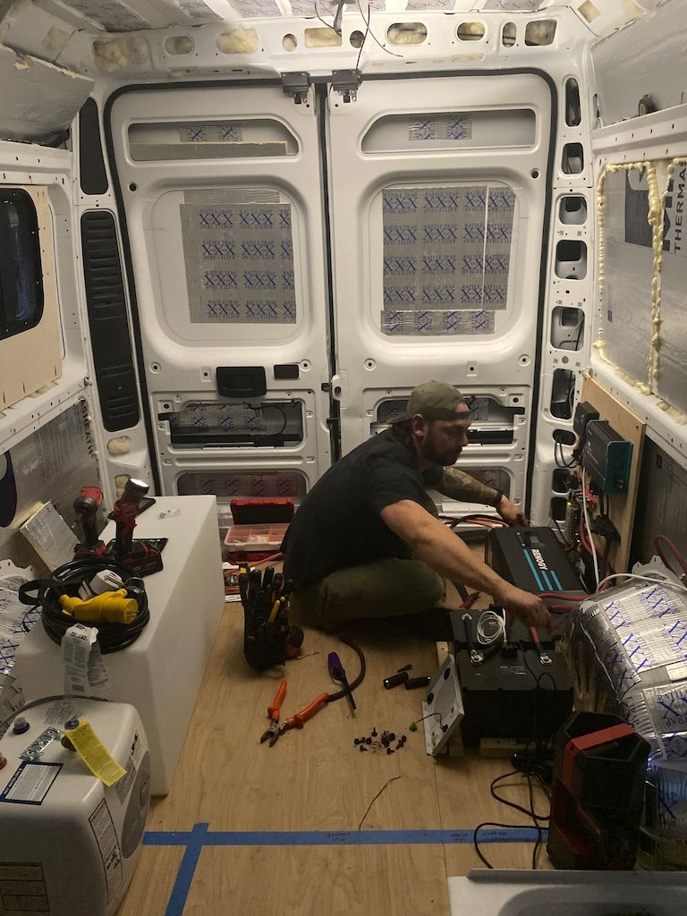 Man working on electrical components inside a van. Tools and materials are scattered around the interior.
