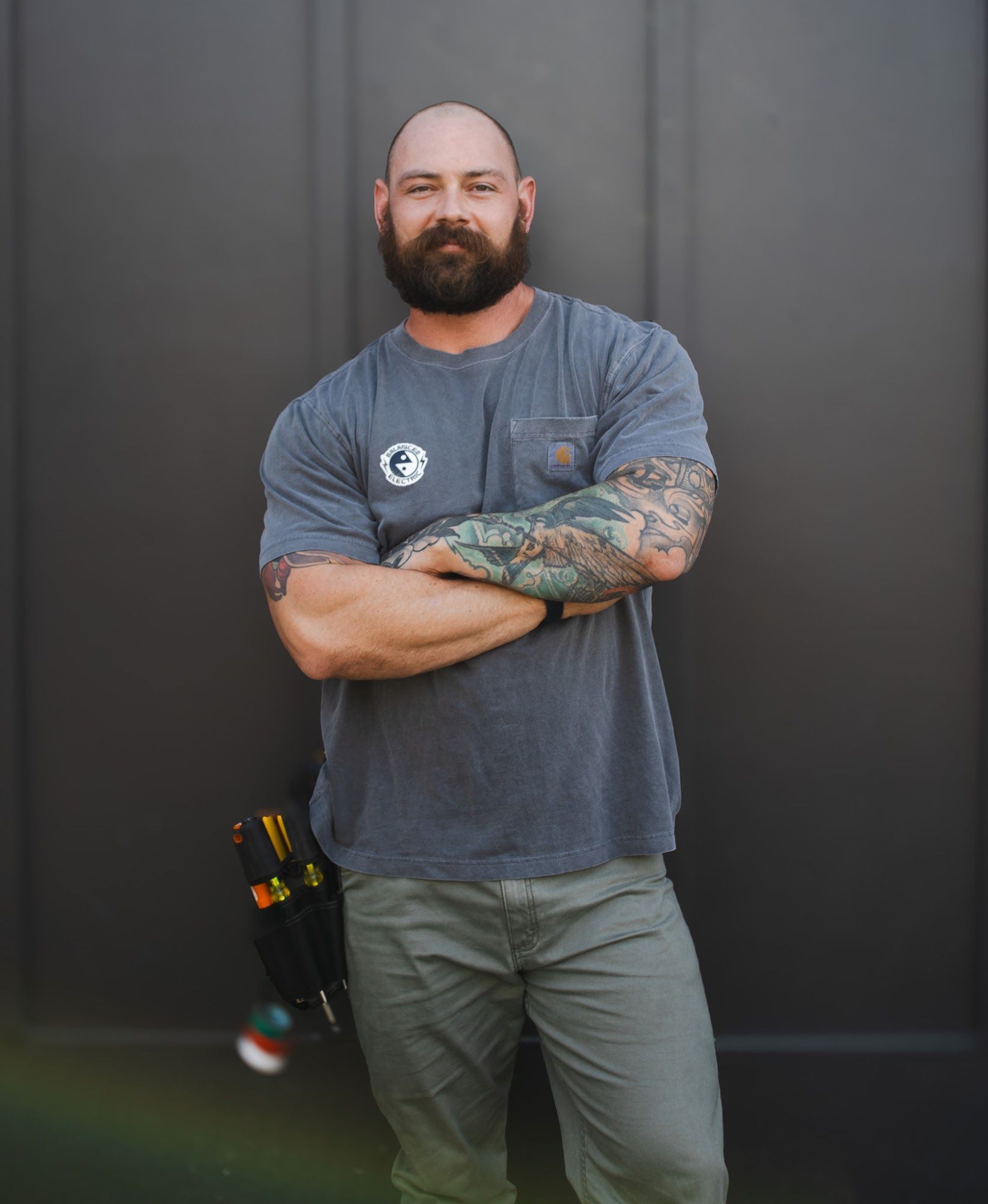 Man with beard and tattoos, arms crossed, wearing gray tee and khaki pants, standing by a dark wall.