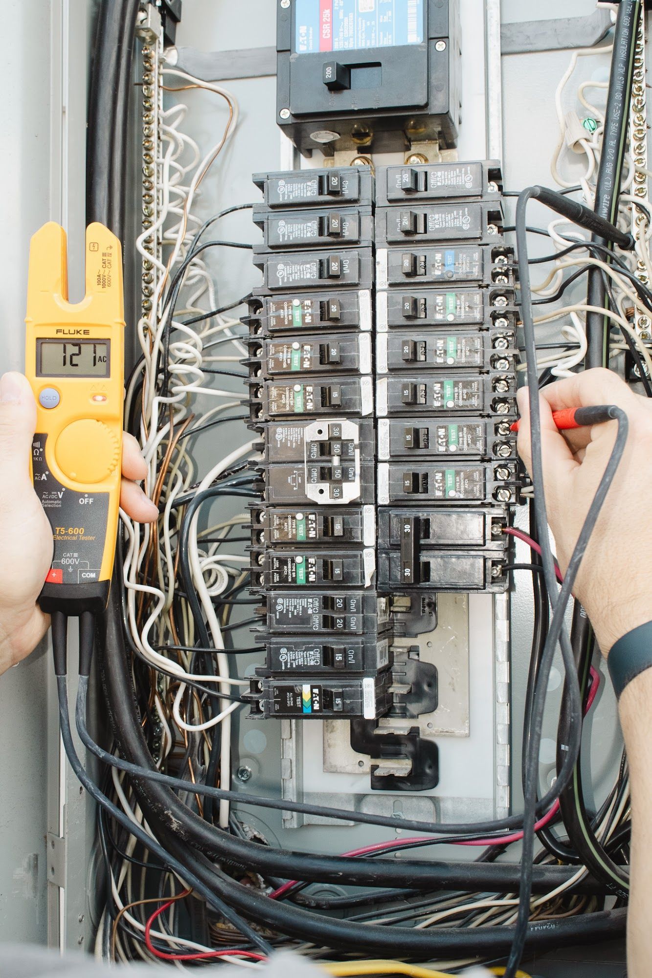 Electrician tests a circuit breaker panel with a multimeter, inside a metal electrical panel.