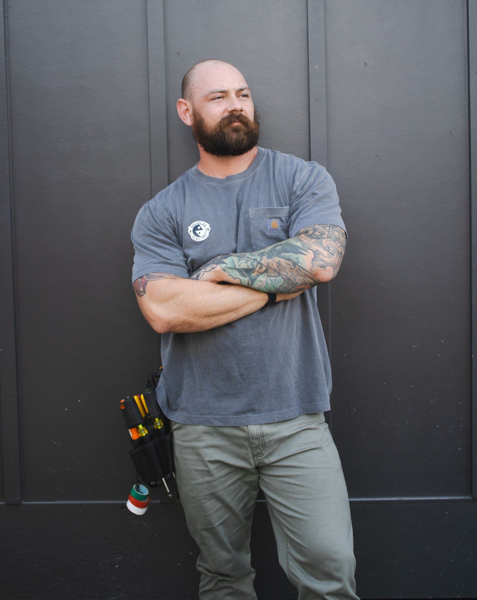 Man with tattoos, beard, and tool belt leaning against a dark door, arms crossed, looking away.