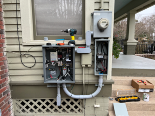 Exterior electrical panel installation on a house with open boxes, tools, and conduit.