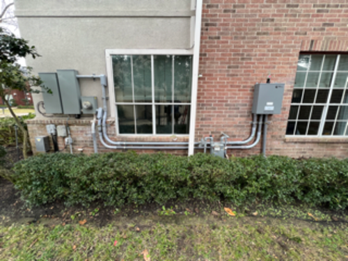 Exterior view of a brick building with electrical boxes, conduits, and windows above green bushes.
