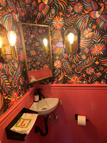 Small bathroom with floral wallpaper, pink accents, gold mirror, and sink.