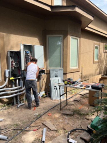 Construction worker installing electrical equipment on a tan house exterior. Wires and tools are on the ground.