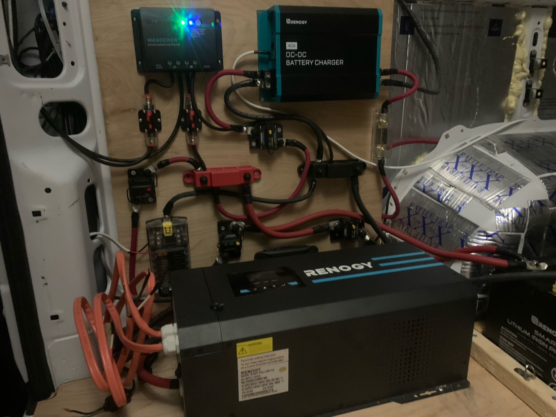 Van electrical system: Inverter, solar charger, DC-DC charger, fuses, wiring. Black, blue, red wires mounted on wood panel.