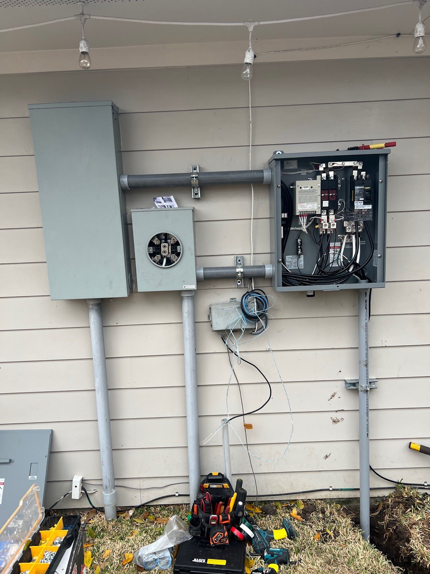 Electrical panel components mounted on a beige wall with gray conduit and wires. Tools and equipment sit at the base.