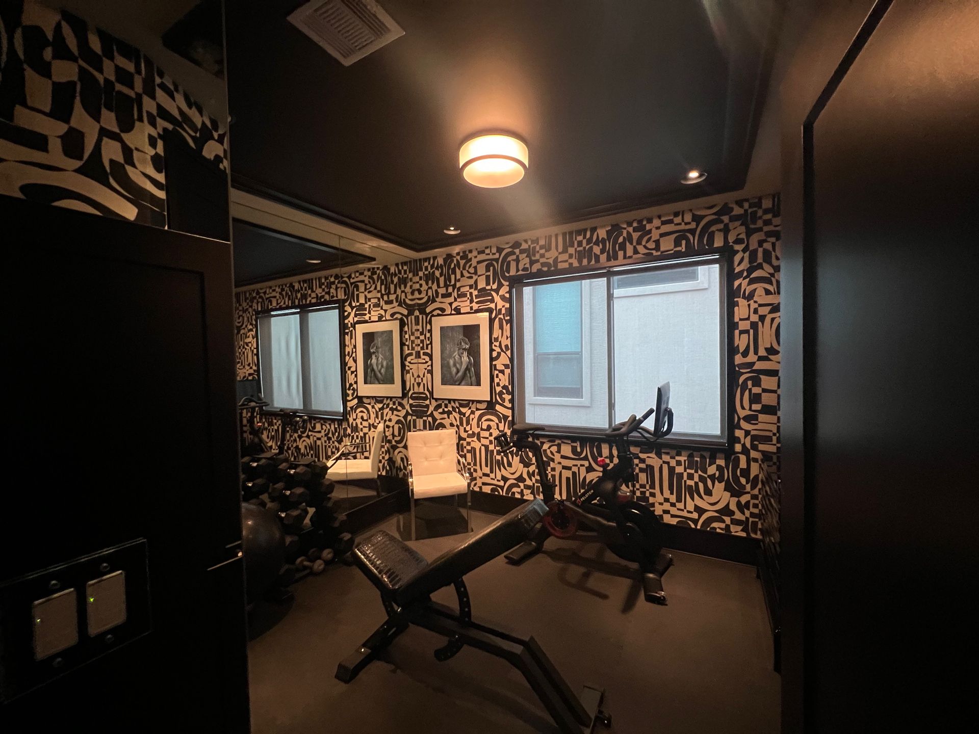 Home gym with black and white patterned wallpaper, weight bench, exercise equipment, and windows.