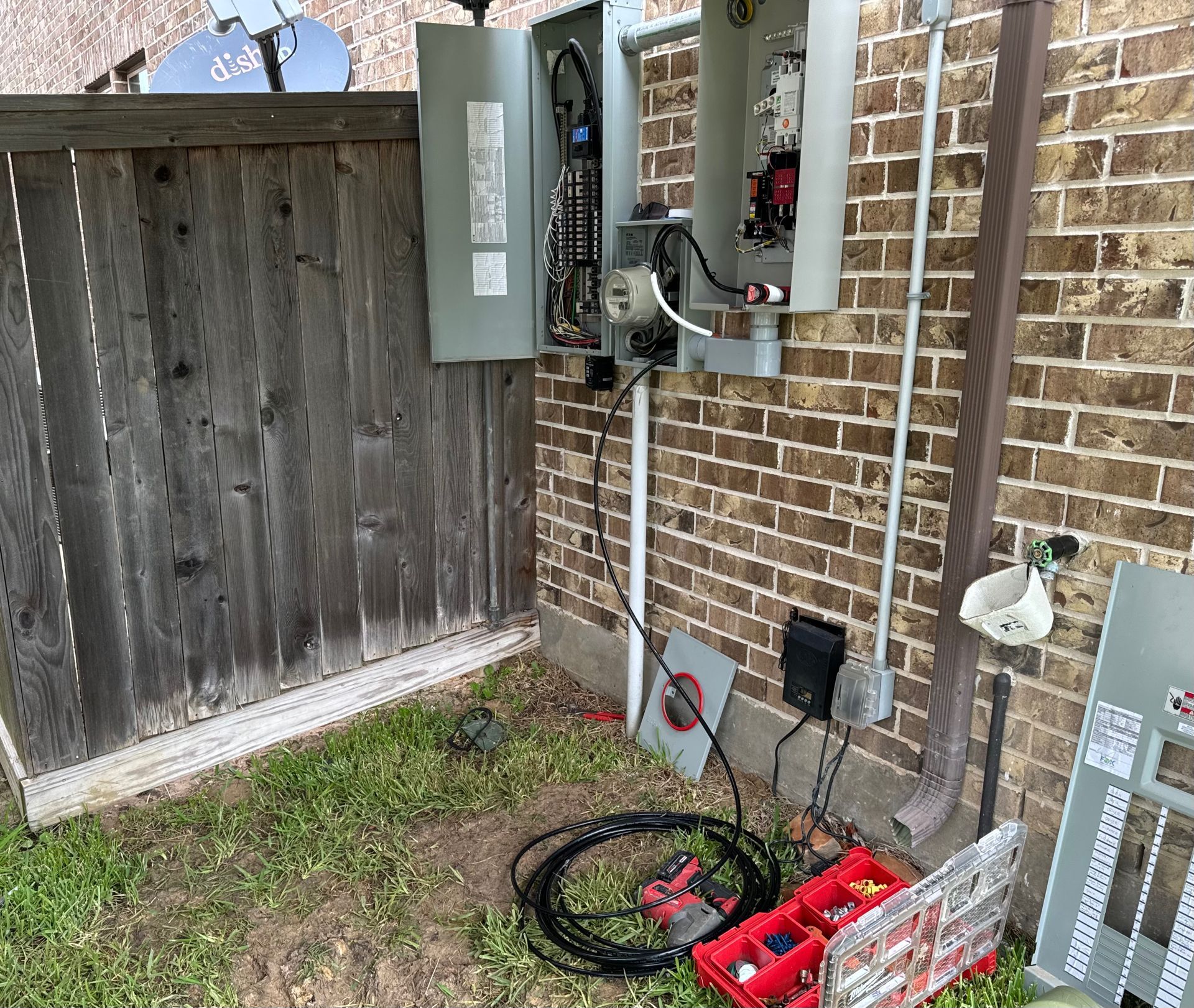 Electrical panel and meter on a brick wall, wires, tools, and a wooden fence.