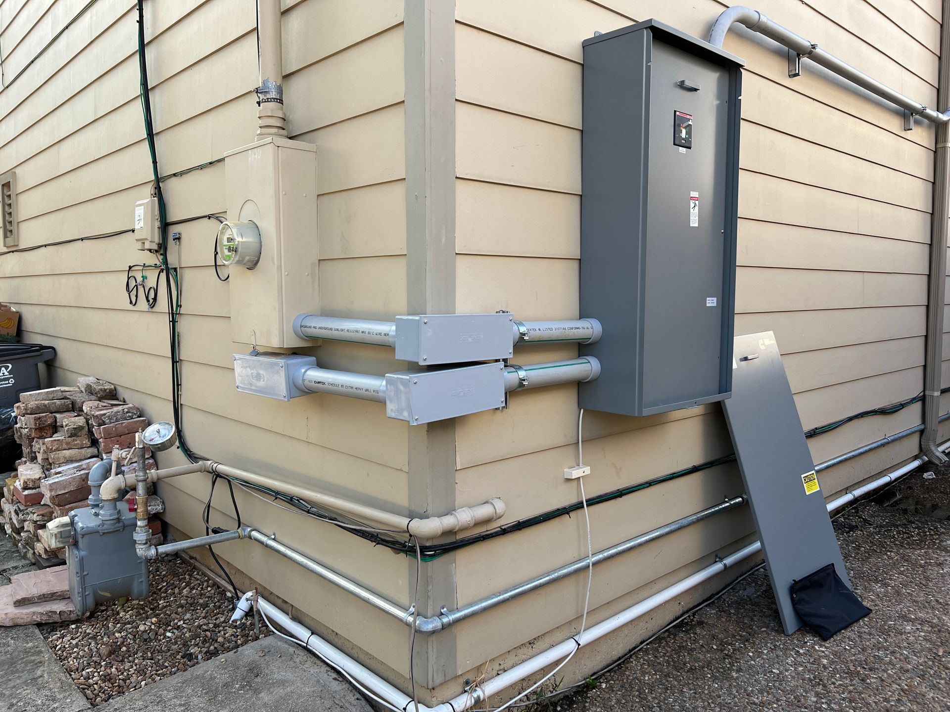 Electrical panel and conduit mounted on a beige building exterior with a gas meter visible.