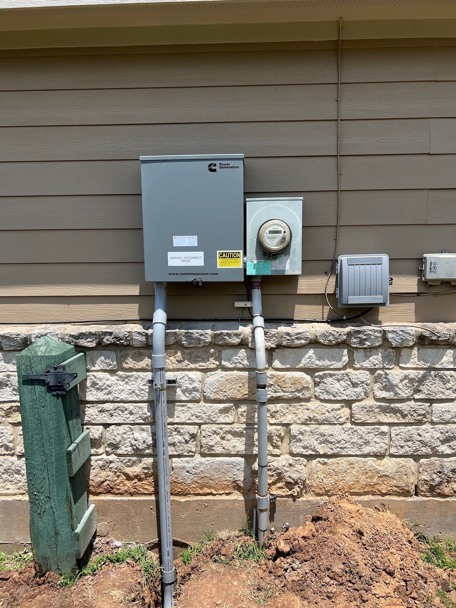 Gray electrical panel and meter on a building's exterior wall, mounted above a brick foundation.