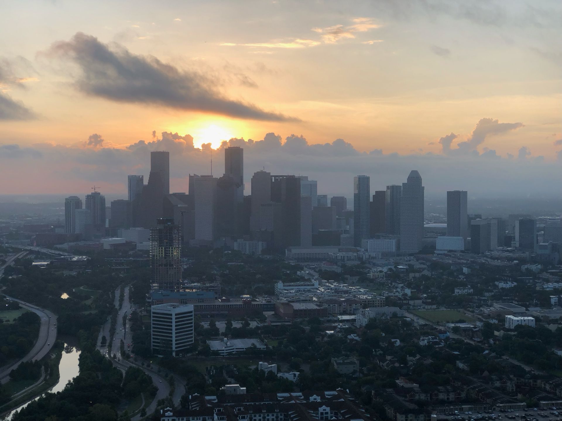 City skyline at sunset, sun behind clouds. Buildings silhouetted, orange and gray sky.