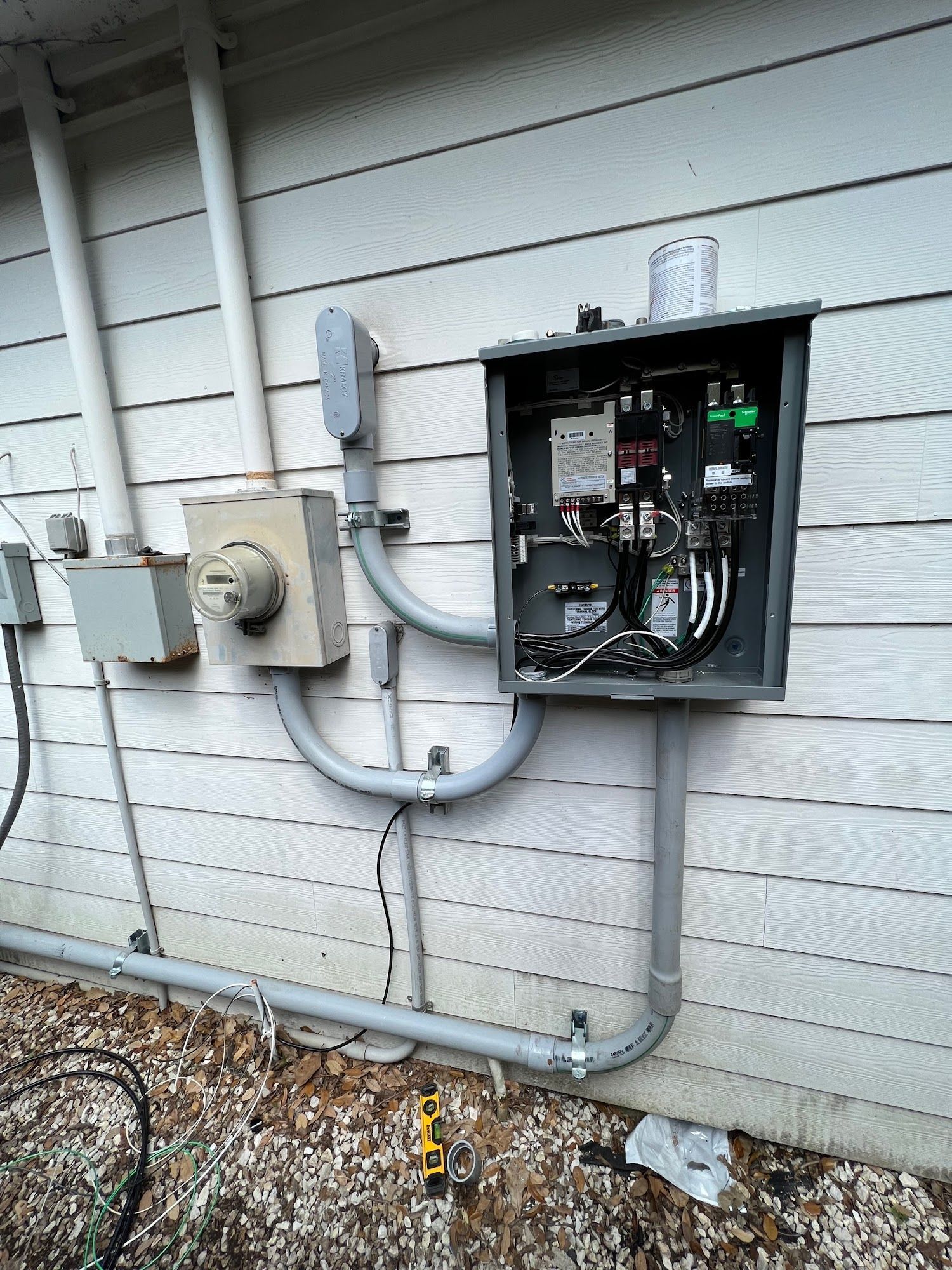 Electrical components mounted on a house's exterior wall: meter, conduit, junction boxes, and open electrical panel.