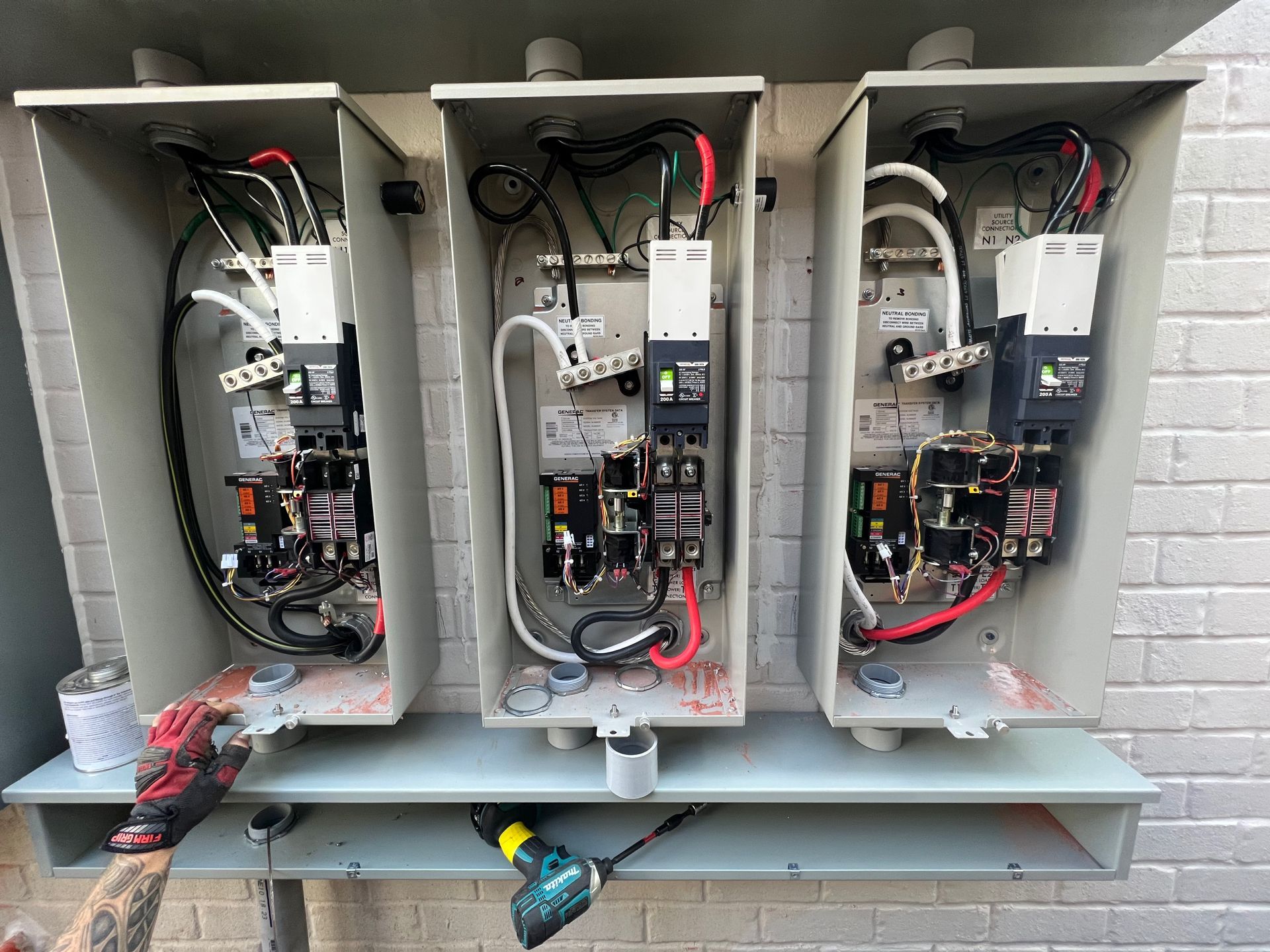 Three electrical control boxes mounted to a wall; a person is working on the bottom left box with a drill.