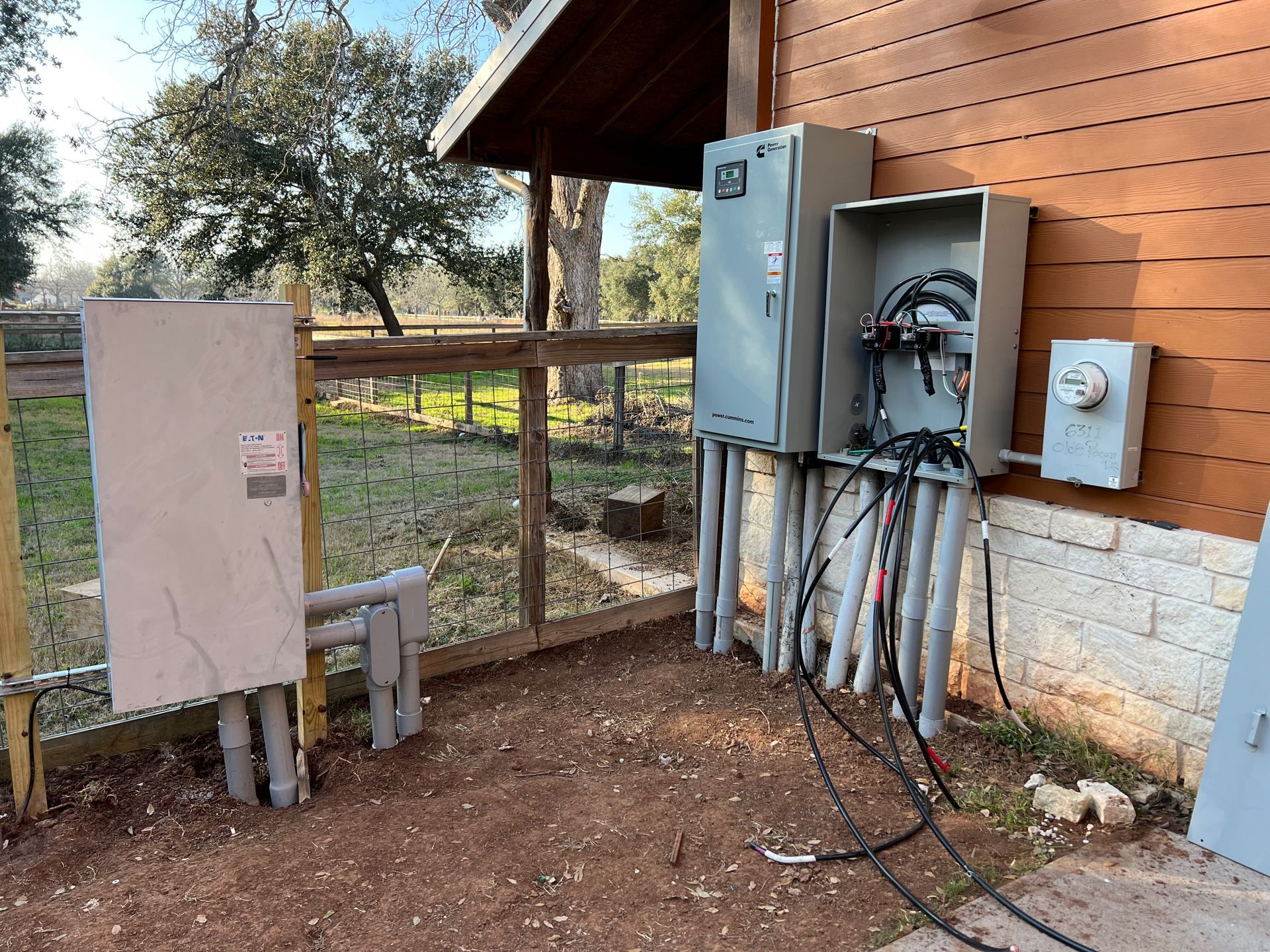 Electrical equipment mounted on a building's exterior with conduit and wiring. Grey boxes, brown building, dirt ground.