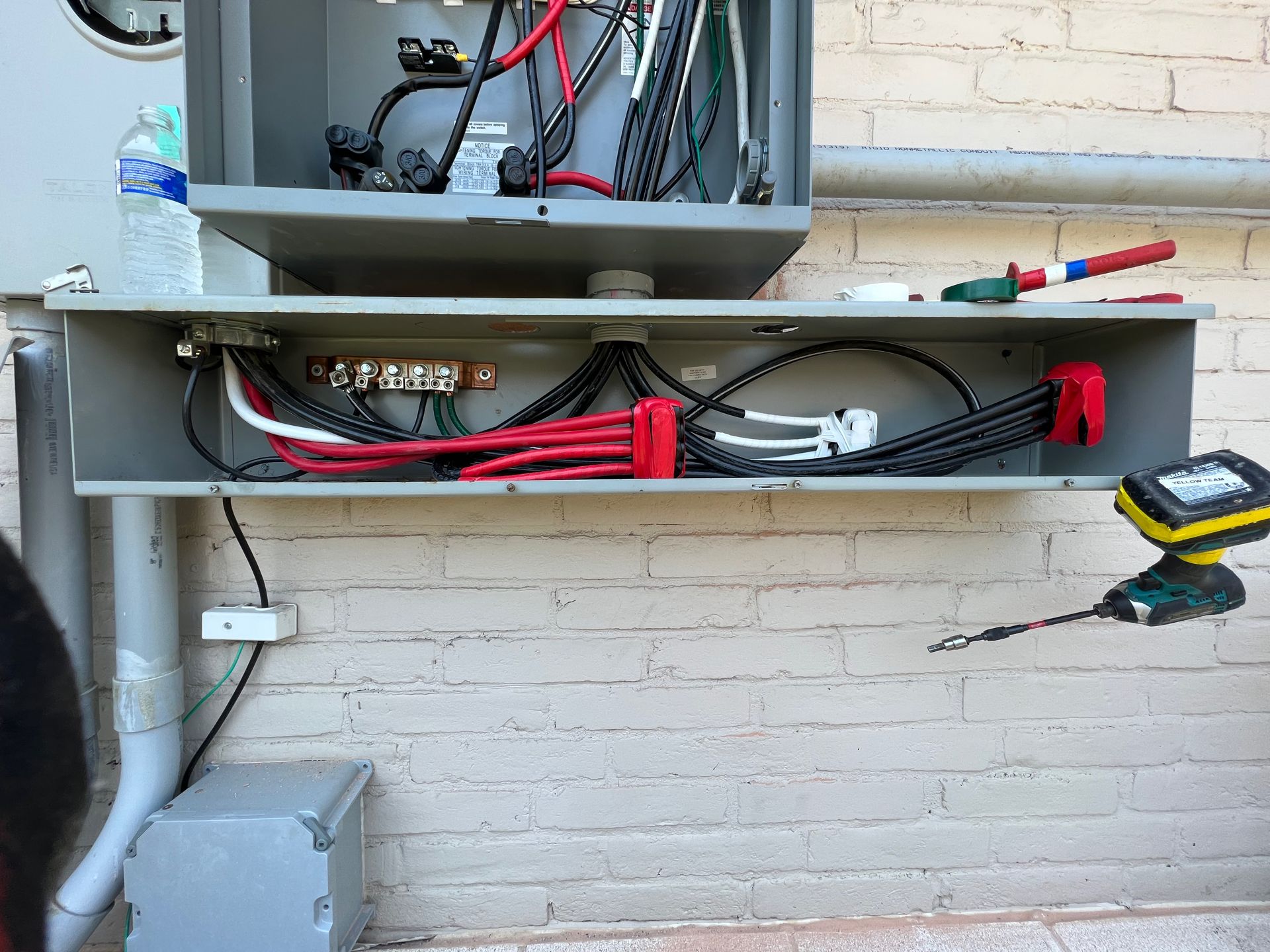 Electrical panel with exposed wires, installed on brick wall, tools nearby.