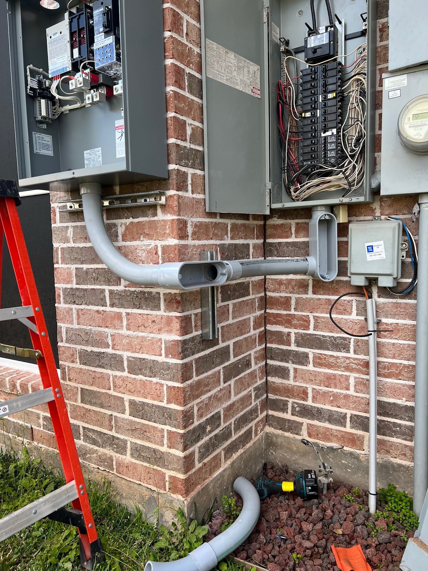 Electrical boxes and conduit on a brick wall, with a ladder and landscaping in the scene.