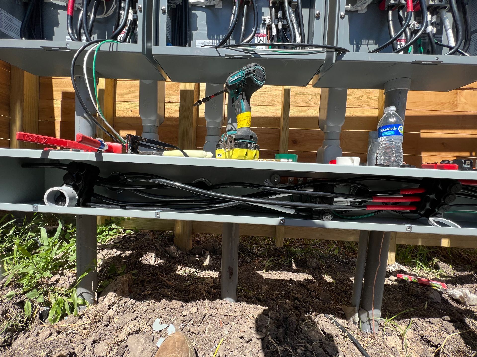 Electrical wiring work in progress outdoors, tools and wires on a gray shelf, wooden fence in the background.