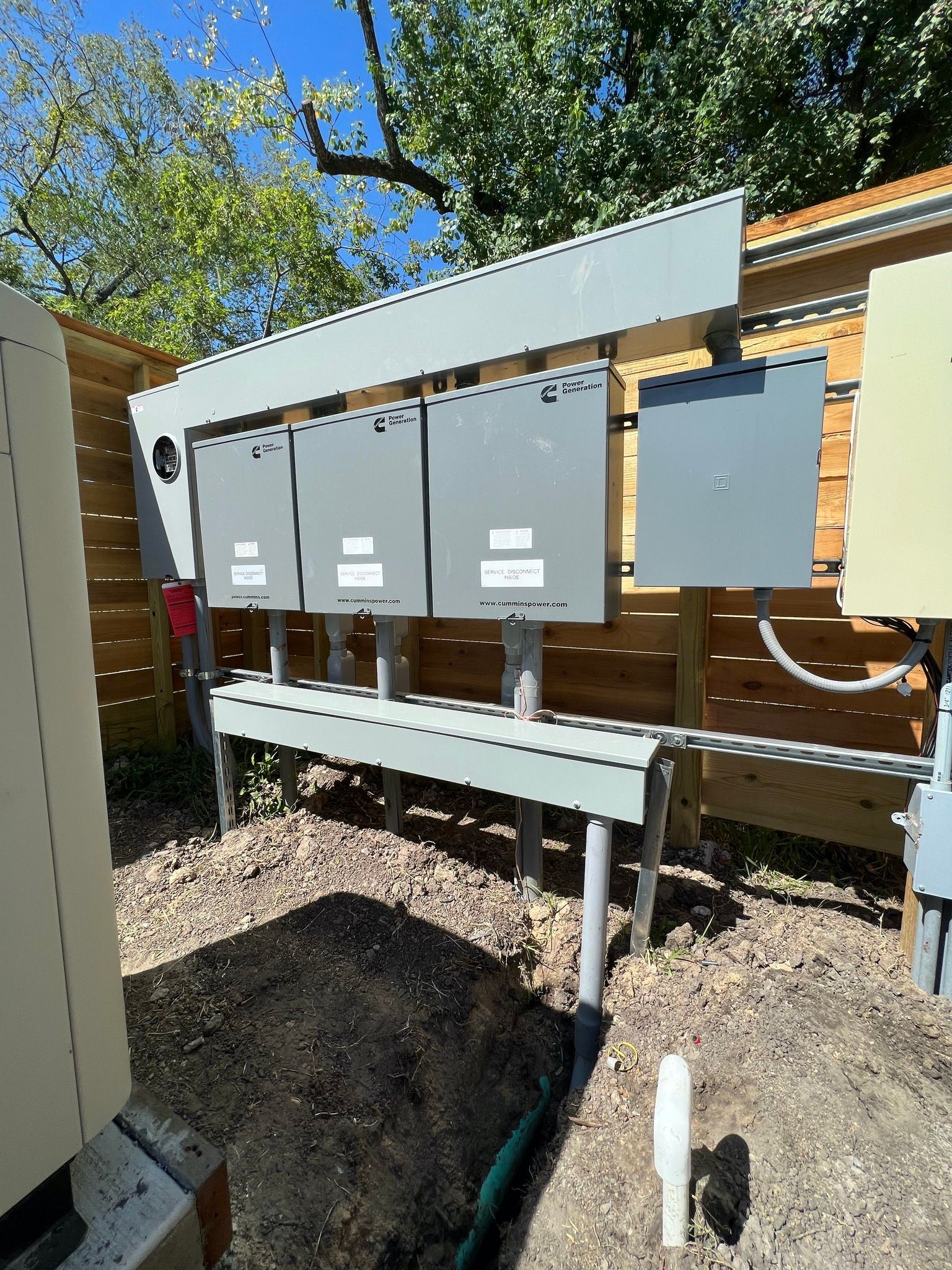 Gray electrical boxes on a metal frame, near a wooden fence and greenery, on a sunny day.