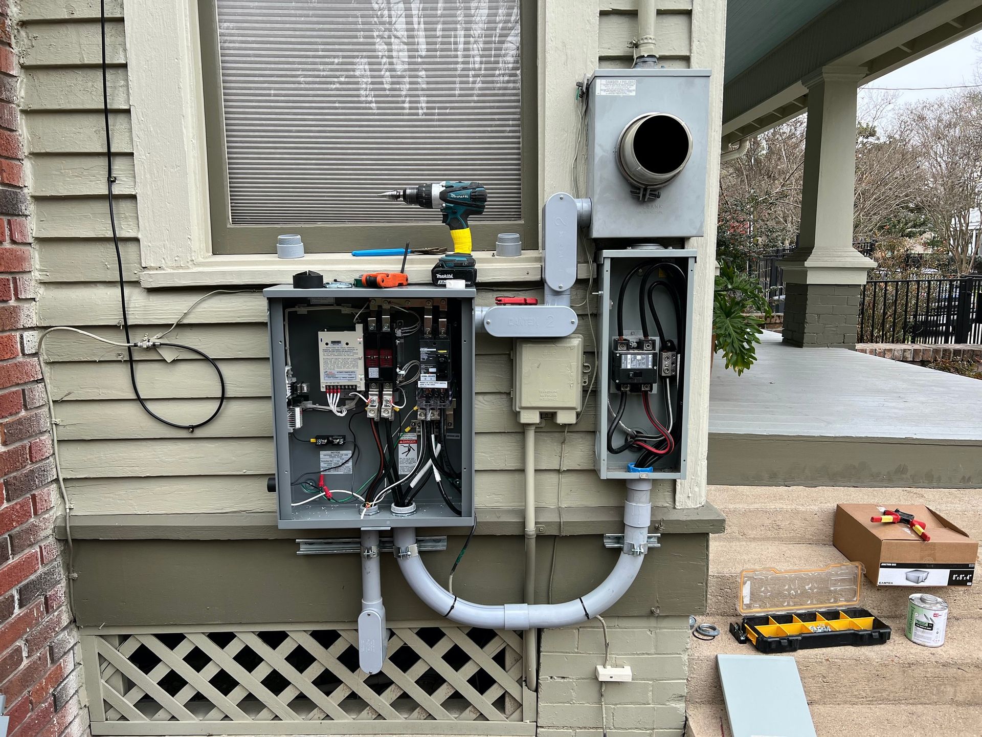 Electrical work on exterior wall; gray metal boxes, wires, drill, and conduit. Beige siding, lattice, and brick.
