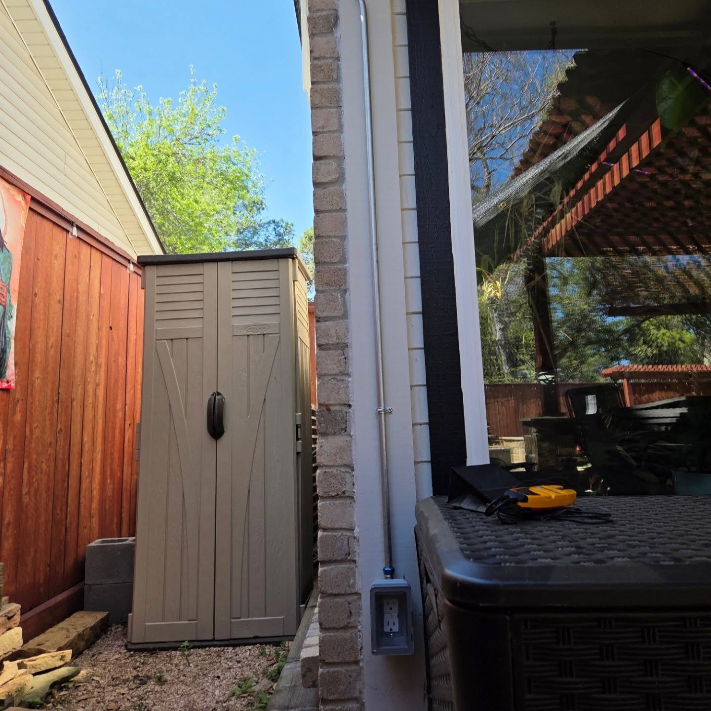 Tan storage shed between a red-stained wooden wall and a brick wall next to a large window.