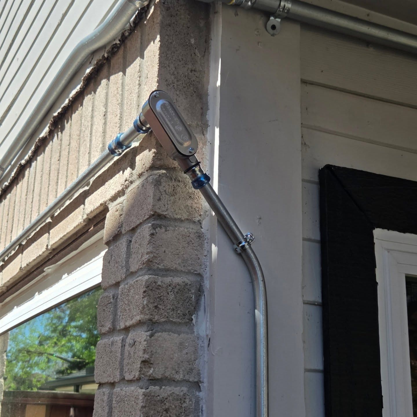 Metal conduit attached to a brick column and siding, leading to an electrical box on an exterior wall.
