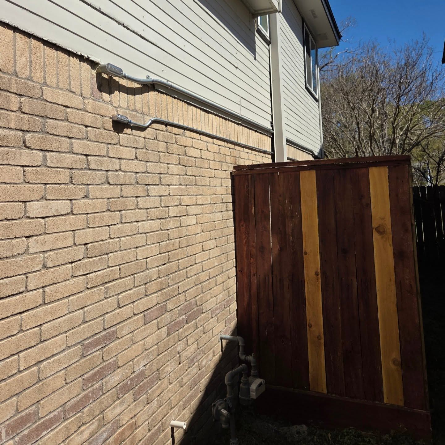 Brick wall of a house with metal conduit. Wooden fence is attached to the house. Blue sky.