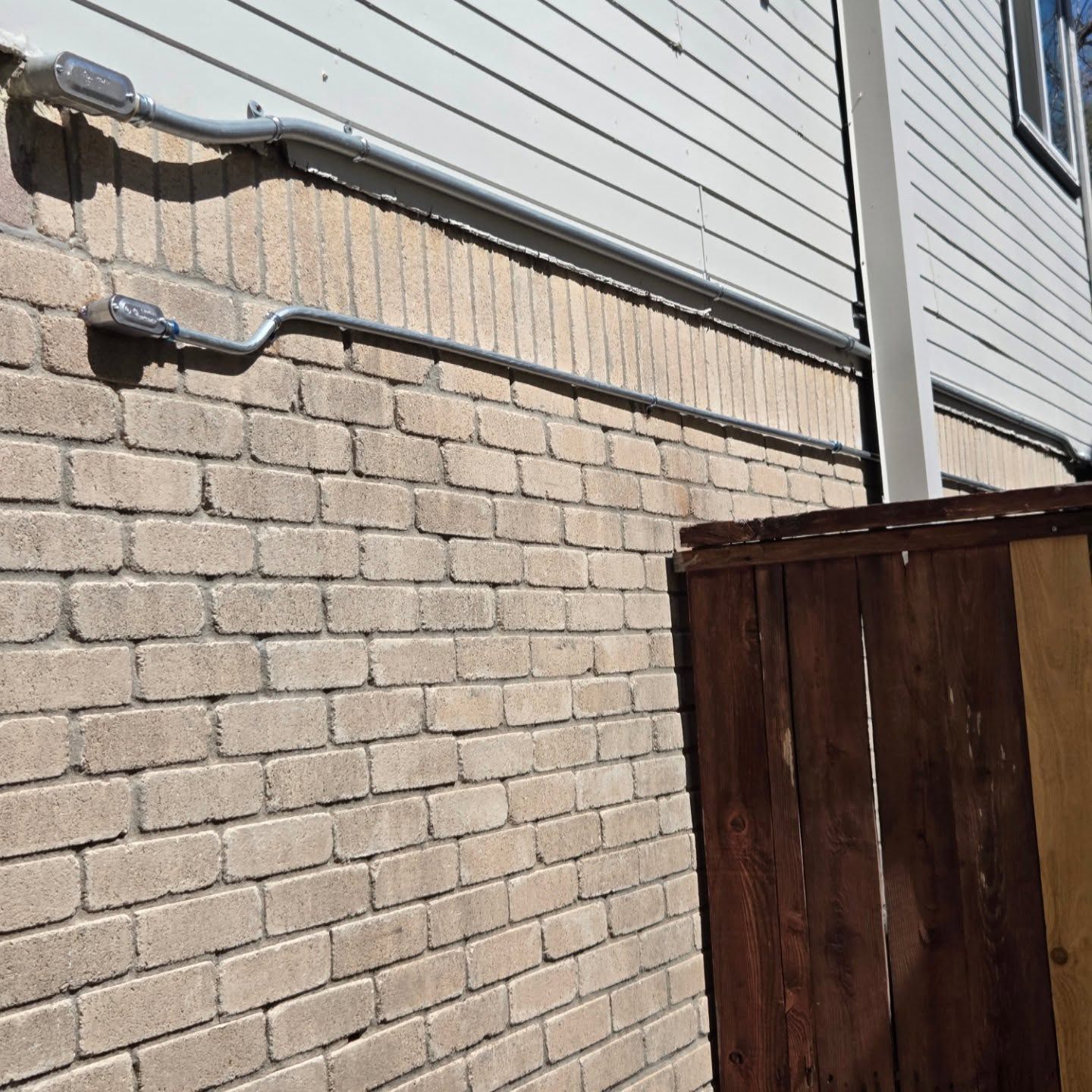 Galvanized electrical conduit running along a brick wall and a white-sided building. Brown wooden fence in foreground.