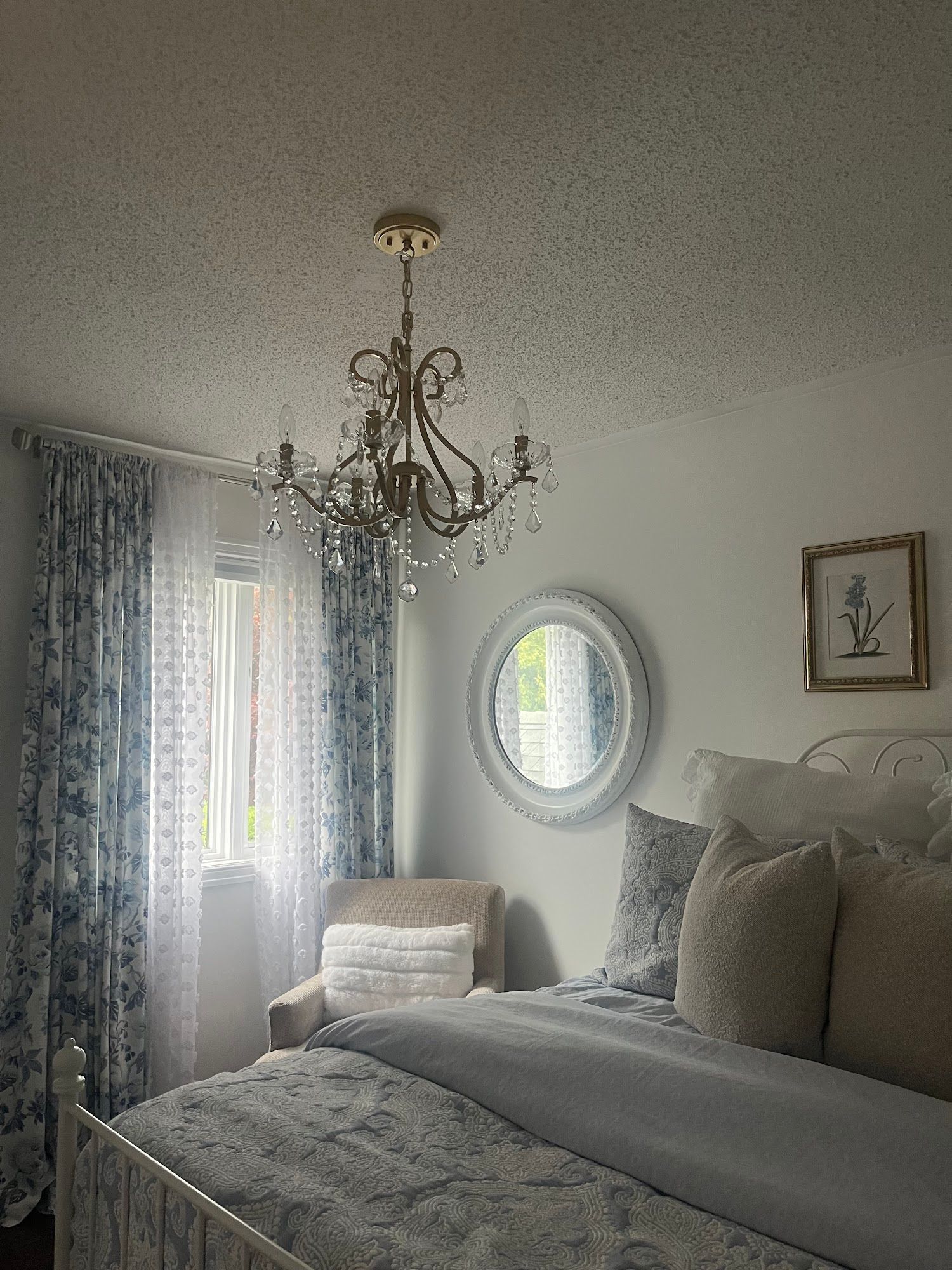 Bedroom with floral curtains, chandelier, round mirror, and bed with pillows.