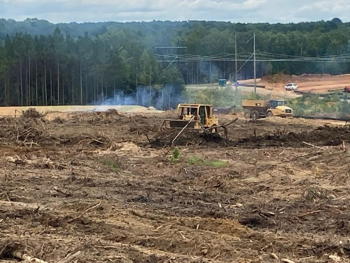 Bulldozers clearing a deforested area for construction. Smoke rises in the background near trees and power lines.